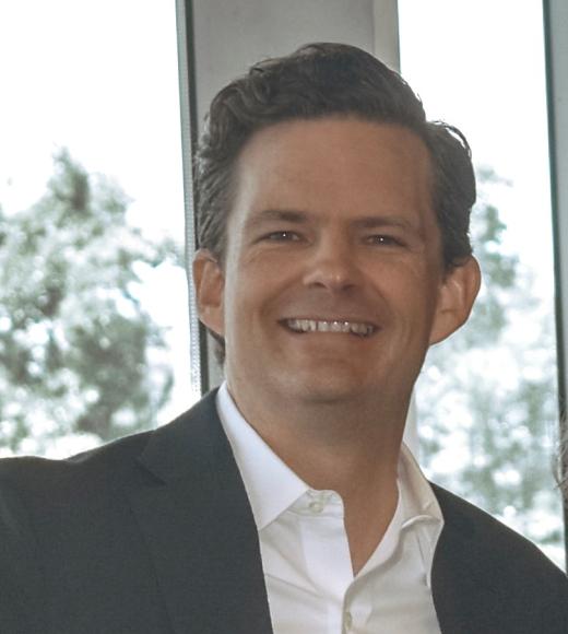 A profile image of a person with short dark hair, wearing a white button down shirt and dark jacket, standing in front of a mostly white background with some greenery.