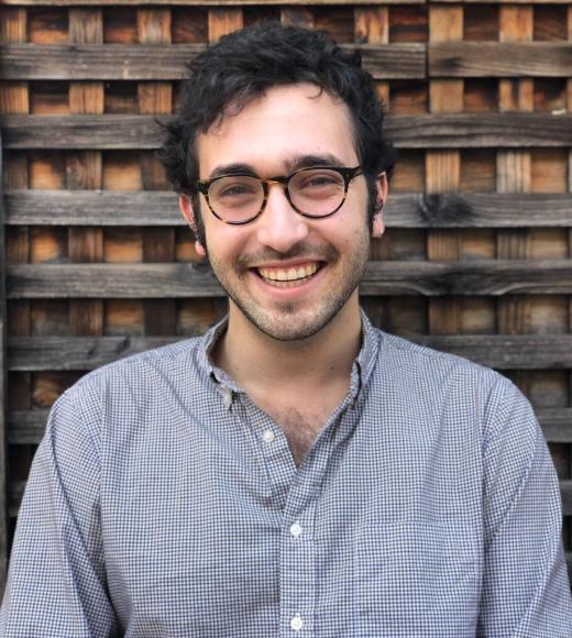 A profile image of a person with short curly hair and glasses, wearing a gray button down shirt and standing in front of a wooden background