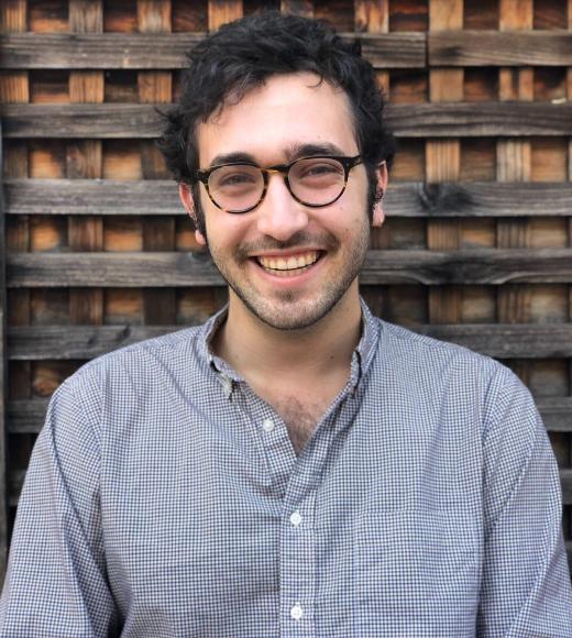 A person with short curly hair and glasses in front of a natural wood background