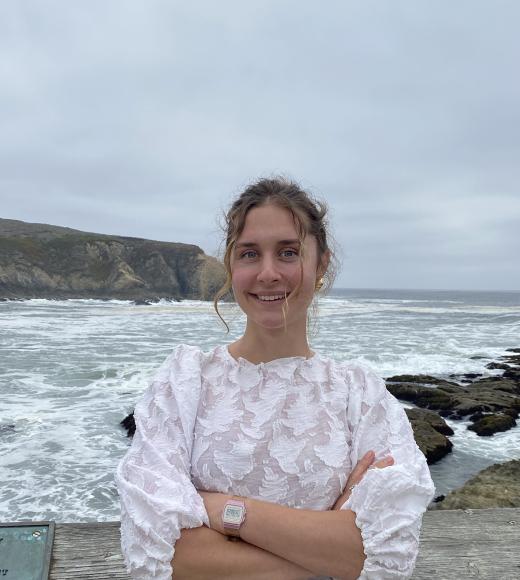 A portrait of Karolina Zabinski, wearing a white shirt and leaning against a wooden structure in front of a beach.