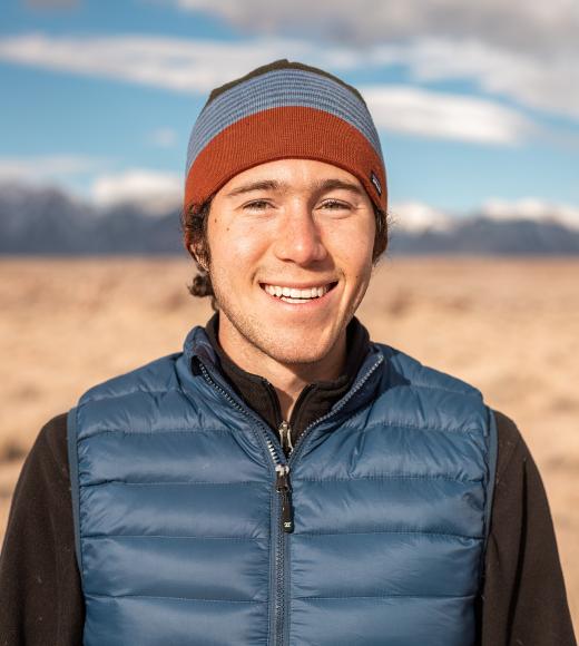 An image of Nicholas Trautman wearing a blue puffer vest and beanie, standing in front of a dry grassy landscape with blue skies and white clouds.