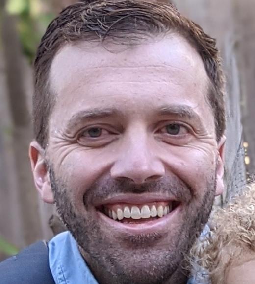 A profile image of a person with short brown hair and facial hair, wearing a blue button down and standing in front of a wooden fence.