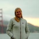 A person in a light colored zip up jacket with medium length blond hair standing in front of the Golden Gate bridge in San Francisco