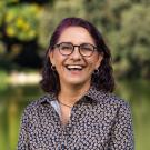 A portrait of Andrea Alfaro standing in front of greenery and joyfully laughing into the camera.