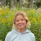 A portrait of Grace Callahan standing in front of a wooded area with yellow flowers.
