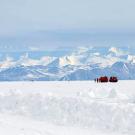 Snowy ground in the foreground and snow covered mountains in the background, against which two bulky red vehicles are visible far off on the horizon, along with several people standing next to the vehicles.