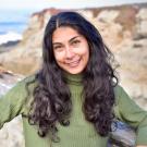 A portrait of Audrey Deutsch standing in front of a coastal background and wearing a high necked green sweater.