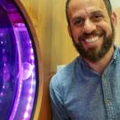 A portrait of Dr. David Gold, smiling and leaning in towards an aquarium full of jellyfish lit with a purple tint.