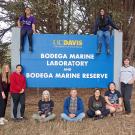 Nine people gathered around a large blue sign that says UC Davis Bodega Marine Laboratory and Bodega Marine Reserve