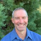 A profile image of a person with short hair wearing a button down blue shirt and standing in front of greenery.