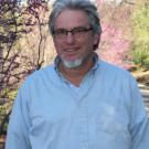 A portrait of John Durand, standing in an outdoor scene wearing a button down blue shirt.