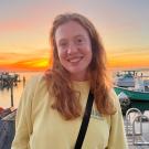 A portrait image of Callie Hundley, standing in front of a sunset lit harbor.