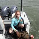 A person kneeling down on a boat next to a mass of marine algae and organisms, giving a thumbs up at the camera.