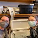 Two people posing in front of a cooler of research equipment smiling with masks on.