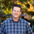 A portrait of Dr. Jay Stachowicz, wearing a dark blue plaid button up shirt and standing in front of a background of blurred trees.