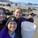 A group of three people gathered on a sandy coast for a selfie, one of them holding up a large white bucket
