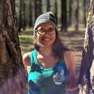 A person in a bright teal tank top and beanie, with long dark hair and black glasses, is smiling at the camera from between two tree trunks.