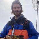 A portrait of Zeke Spooner wearing orange boating waders and holding a fish