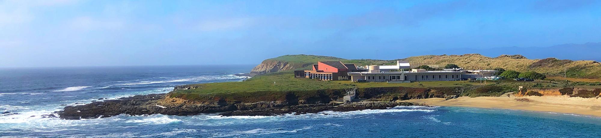 View of Bodega Marine Laboratory with green grass visible in the foreground and the cove in front of the lab building, which is predominantly white with blue skies behind.