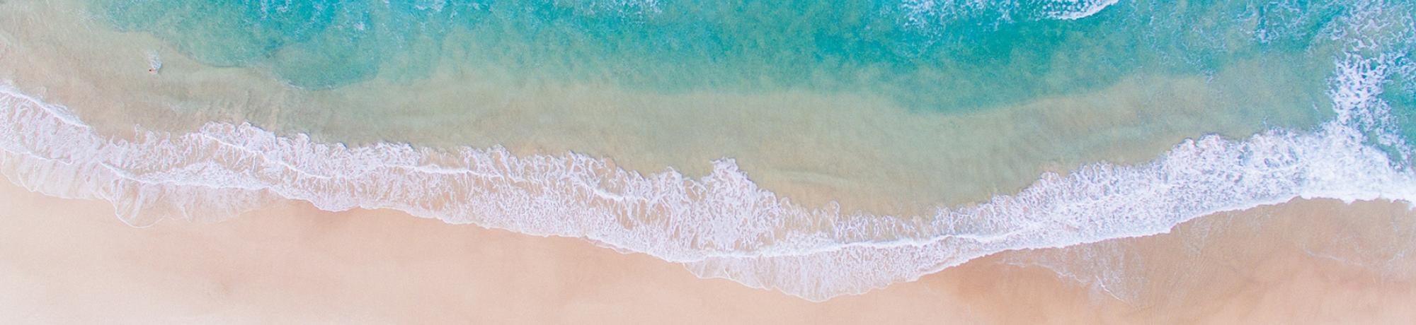 Top down view of teal waves sweeping across a light pink, sandy shore